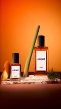 Two bottles of Lush 'Karma' perfume stand on textured stone slabs against an orange background. They are styled with a curl of orange peel, a green stalk of lemongrass, and a small sprig of pine, reflecting the scent's ingredients.